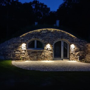 Revonia underground eco-house exterior at night – stone-clad earth-sheltered home with arched windows and green roof